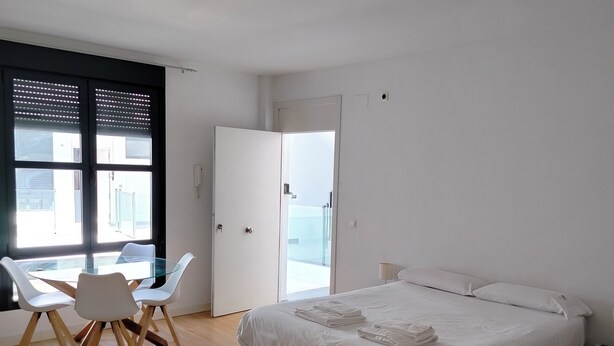 A modern bedroom with a wooden floor, a glass dining table with chairs, a bed with white linens, and a large window with blinds.