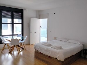 A modern bedroom with a wooden floor, a glass dining table with chairs, a bed with white linens, and a large window with blinds. - Apartamentos Centro Cordoba by MiRa (Córdoba)
