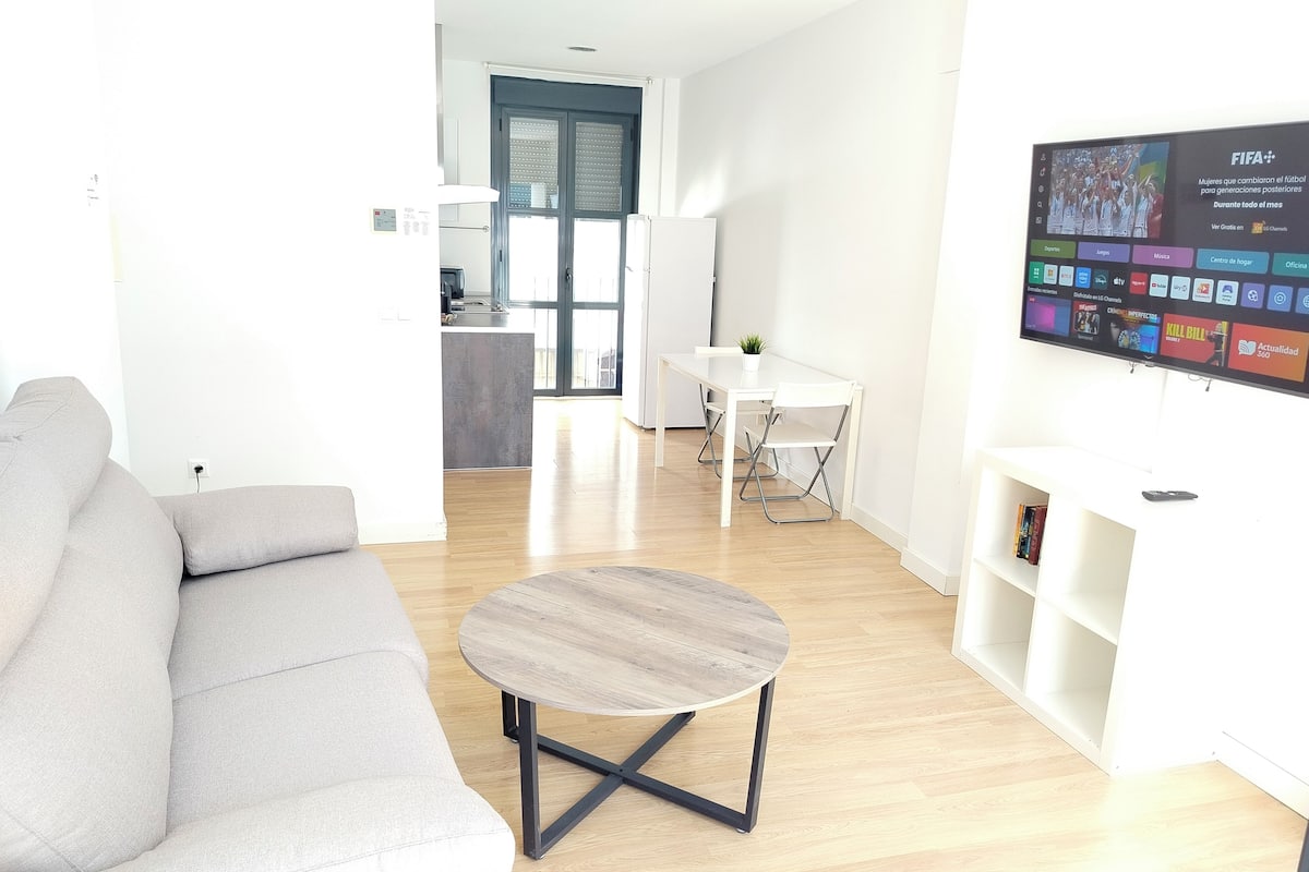 A modern living room with a grey sofa, a round coffee table, and a TV mounted on the wall displaying various channels.