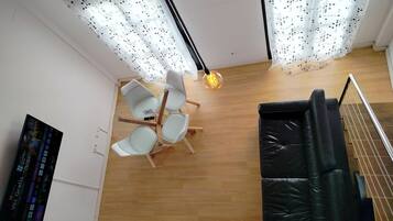 Una habitación con piso de madera, un sofá de cuero negro, una mesa blanca con sillas y una lámpara colgante.
