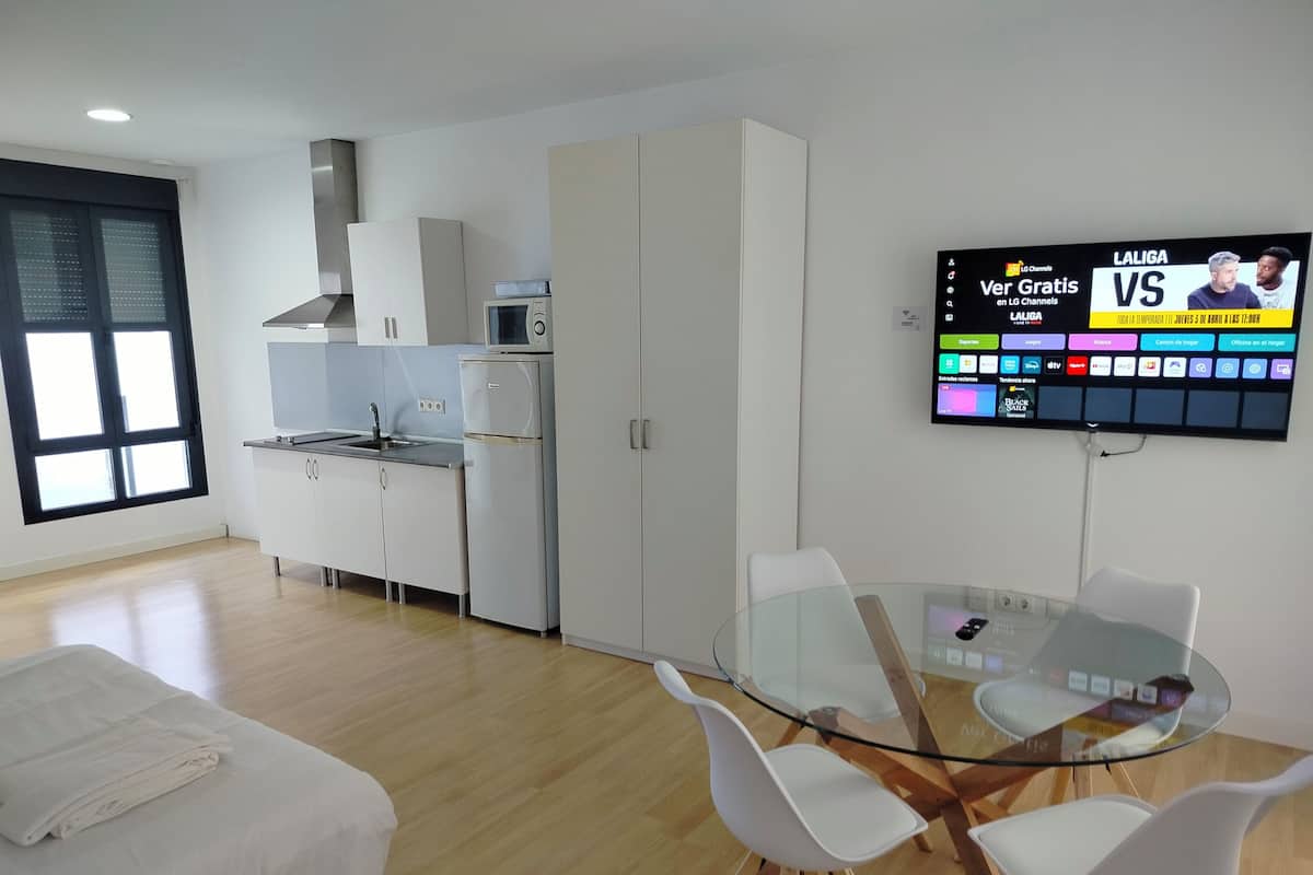 A modern kitchen with white cabinets, a microwave, and a sink. There's a glass table with chairs and a TV displaying a sports channel.