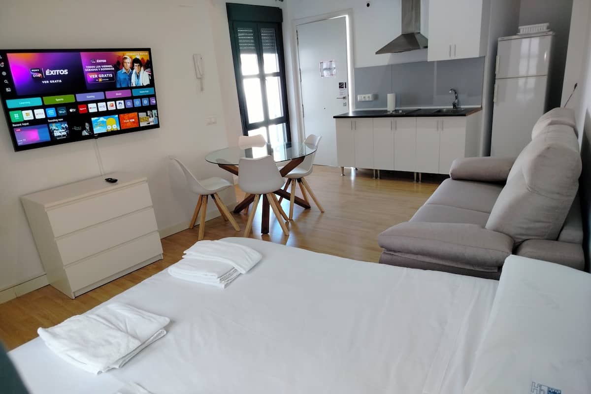 A modern hotel room with a bed, a TV displaying a streaming service interface, a dining area with a table and chairs, and a kitchenette.