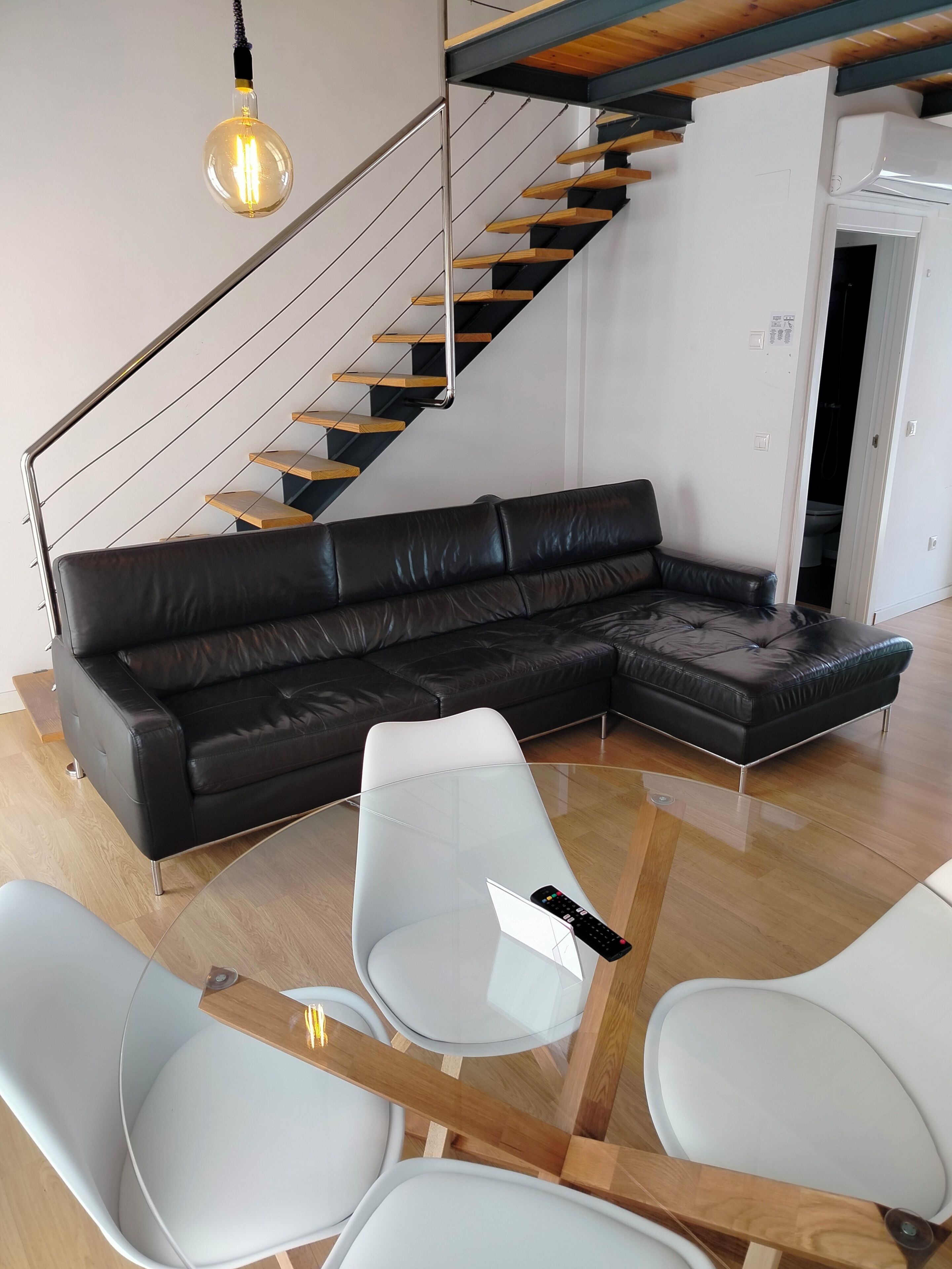 Una sala de estar moderna con un sofá seccional de cuero negro, una mesa de centro de vidrio y sillas blancas. Una escalera de madera con barandilla de metal conduce a un nivel superior.