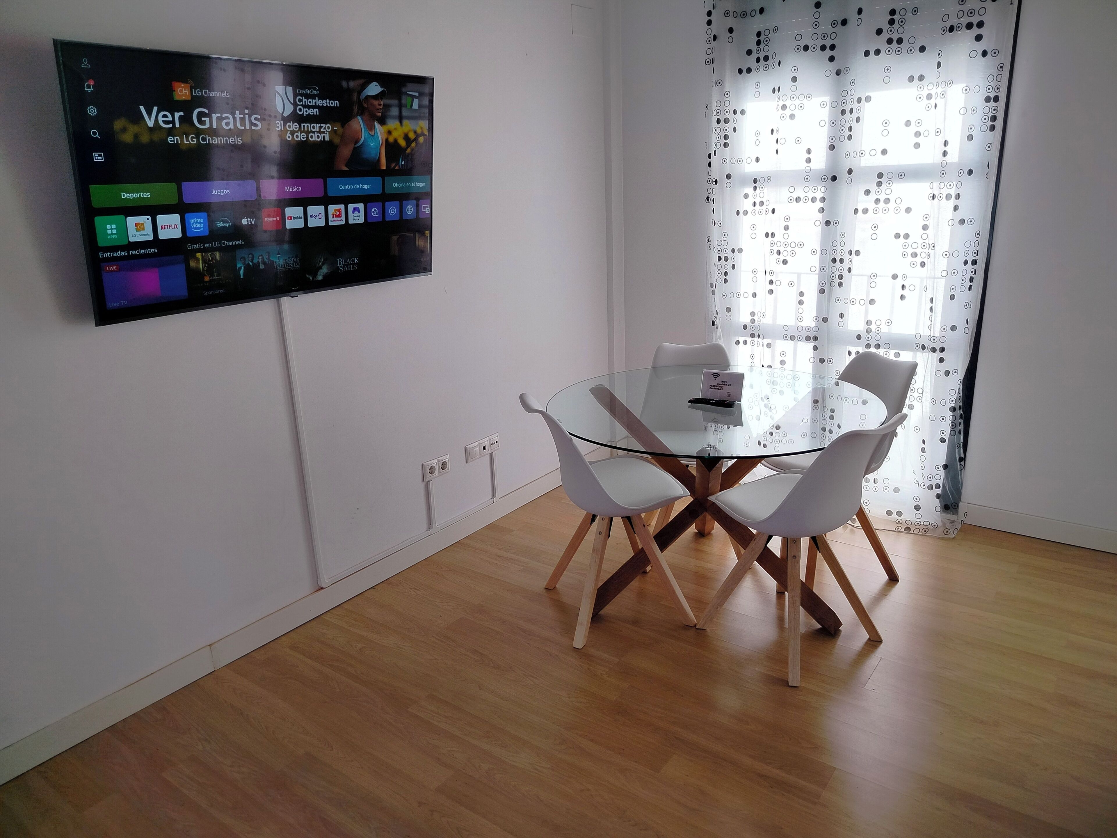 Una camera moderna con una TV a schermo piatto montata a parete, che mostra diversi canali e un evento sportivo in diretta. Si nota un tavolo in vetro con quattro sedie bianche e una piccola lampada da tavolo. La stanza ha il pavimento in legno e una finestra con tenda decorata.