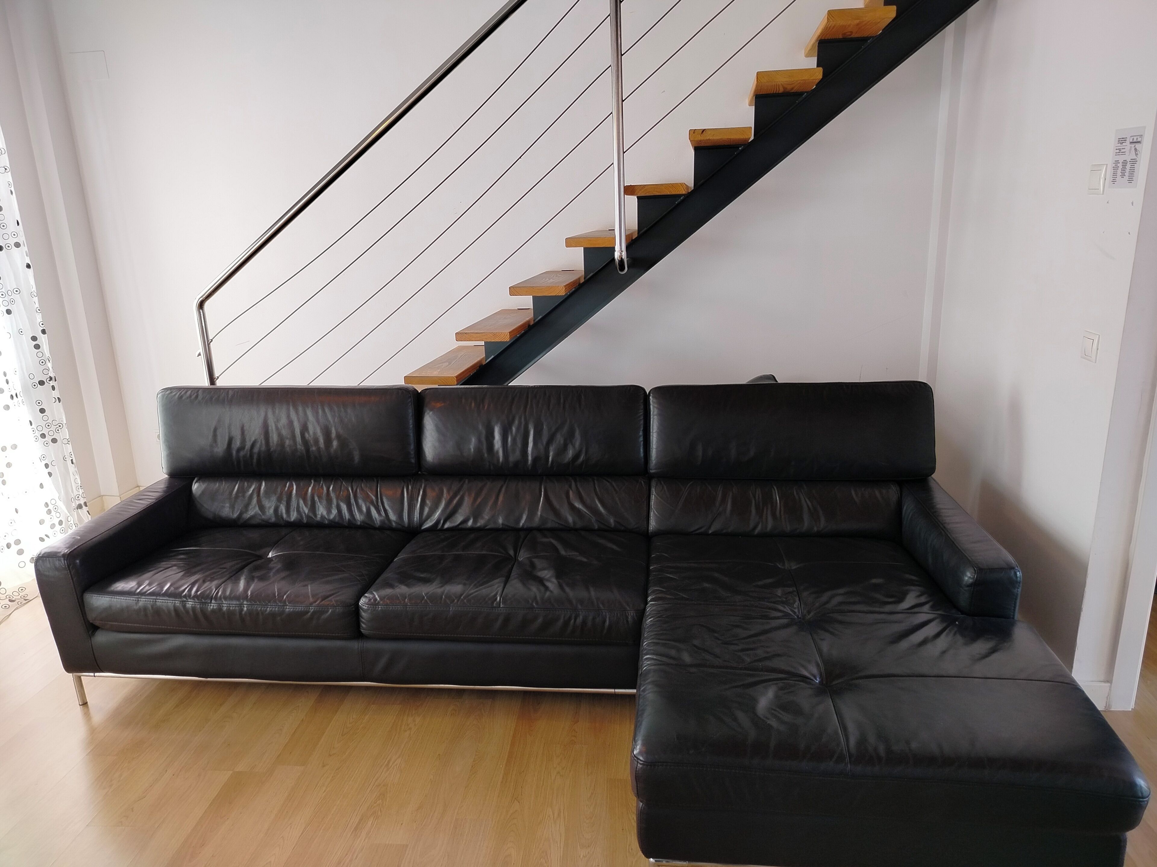 A black leather sectional sofa in a room with a wooden staircase and light-colored wooden flooring.