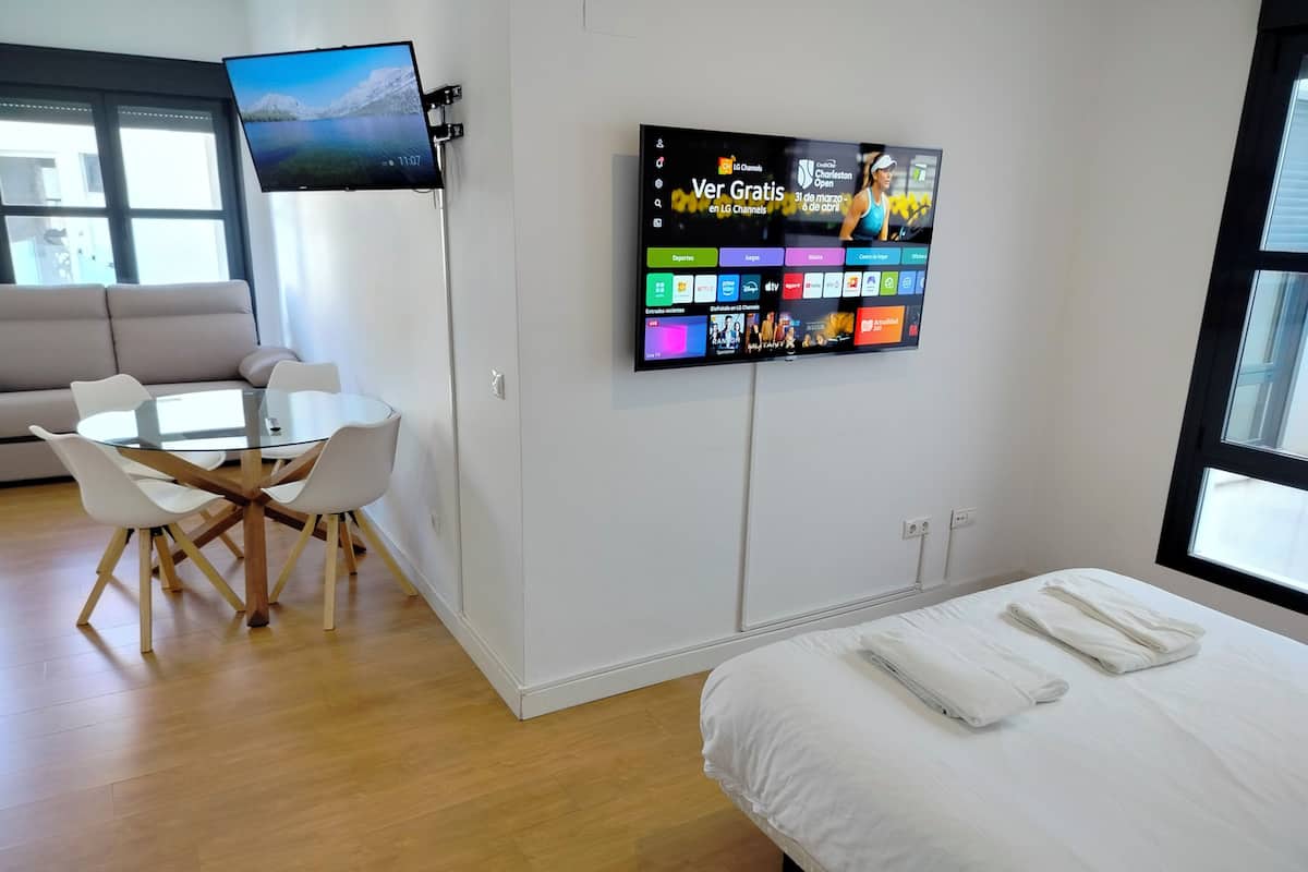 A modern hotel room with a flat-screen TV mounted on the wall displaying various channels, a round glass table with white chairs, a neatly made bed, and a large window providing natural light.