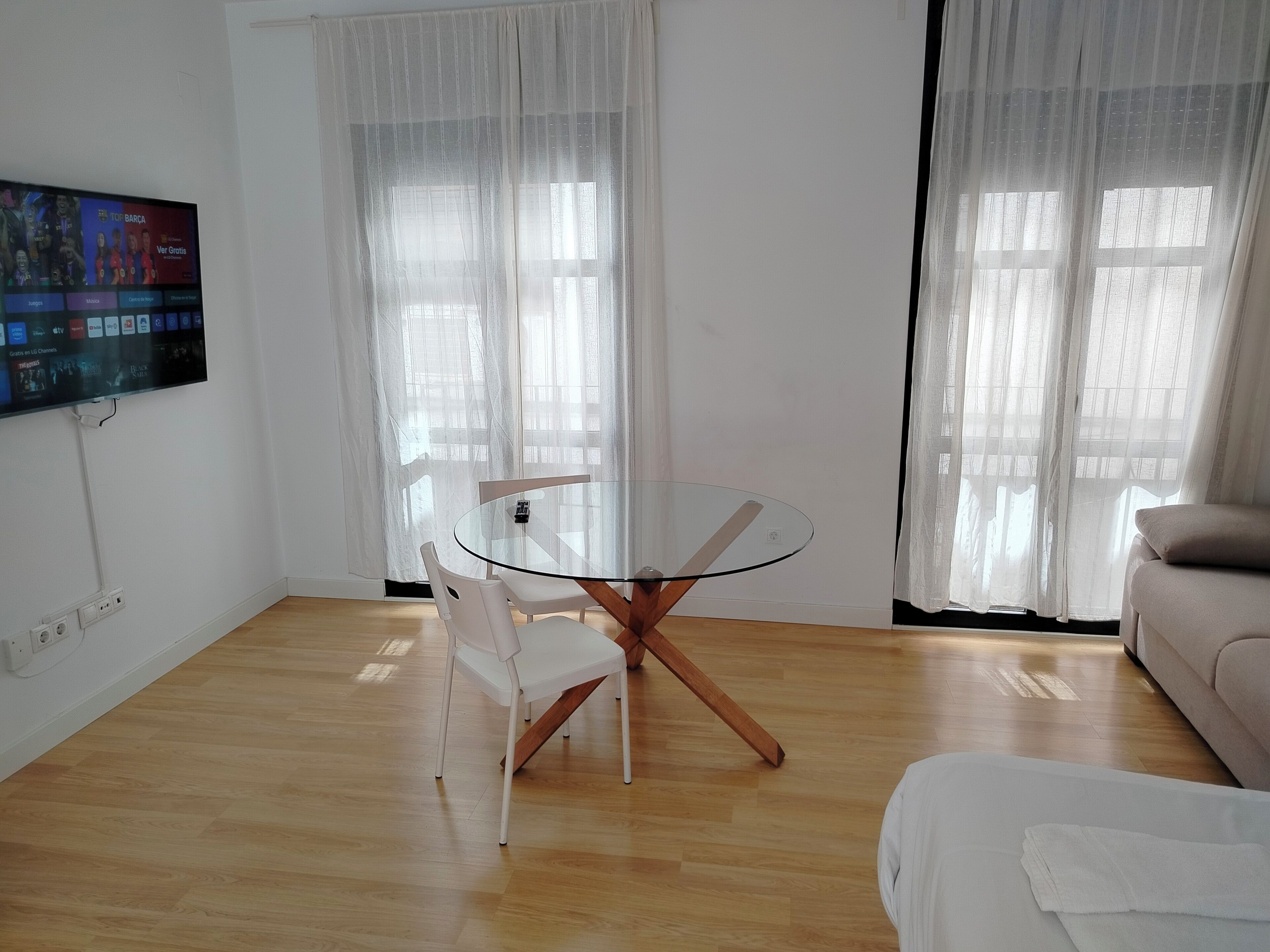 Una sala moderna con una mesa de comedor de cristal, sillas blancas, un sofá beige y un televisor de pantalla plana montado en la pared.