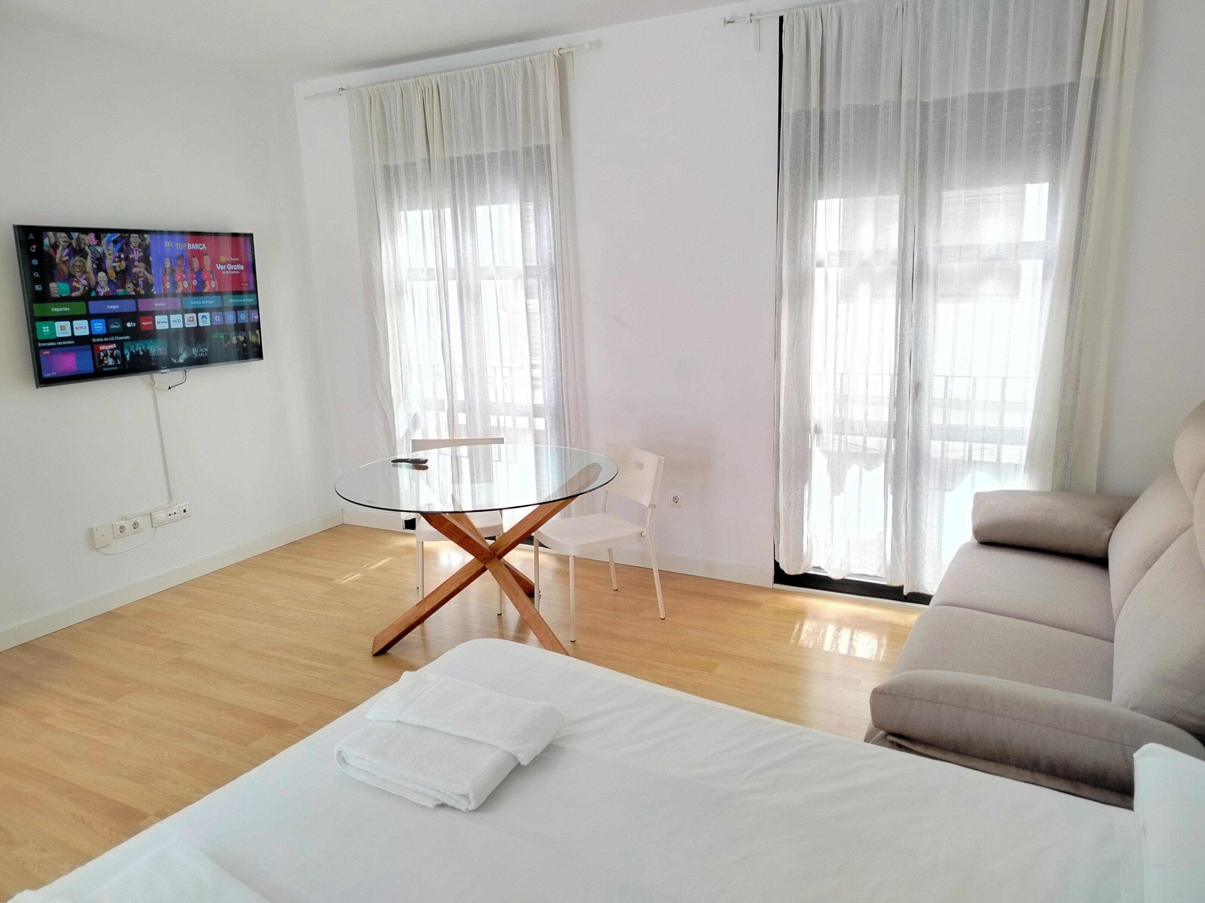 A modern hotel room with a glass table, white chairs, a sofa, and a TV displaying a sports channel.