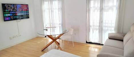 A modern hotel room with a glass table, white chairs, a sofa, and a TV displaying a sports channel.