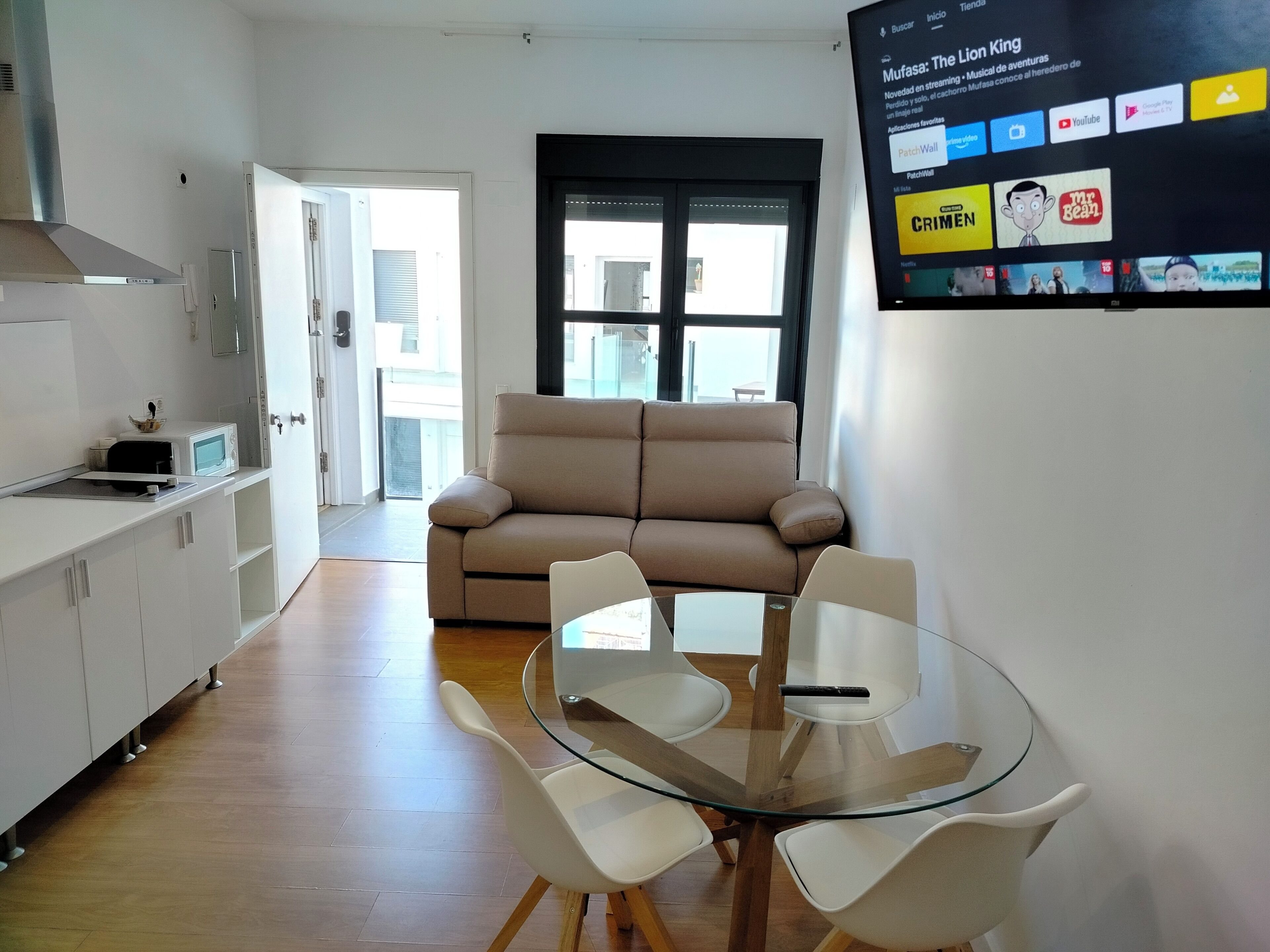 Un salon moderne avec un canapé beige, une table à manger en verre entourée de quatre chaises, un téléviseur à écran plat fixé au mur diffusant un service de streaming, et un coin cuisine doté de meubles blancs et d’un four à micro-ondes.