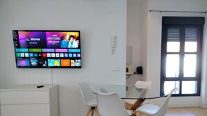 A modern living room with a flat-screen TV mounted on the wall displaying various streaming services. There is a glass-top dining table with white chairs, a white cabinet, and a window with blinds.