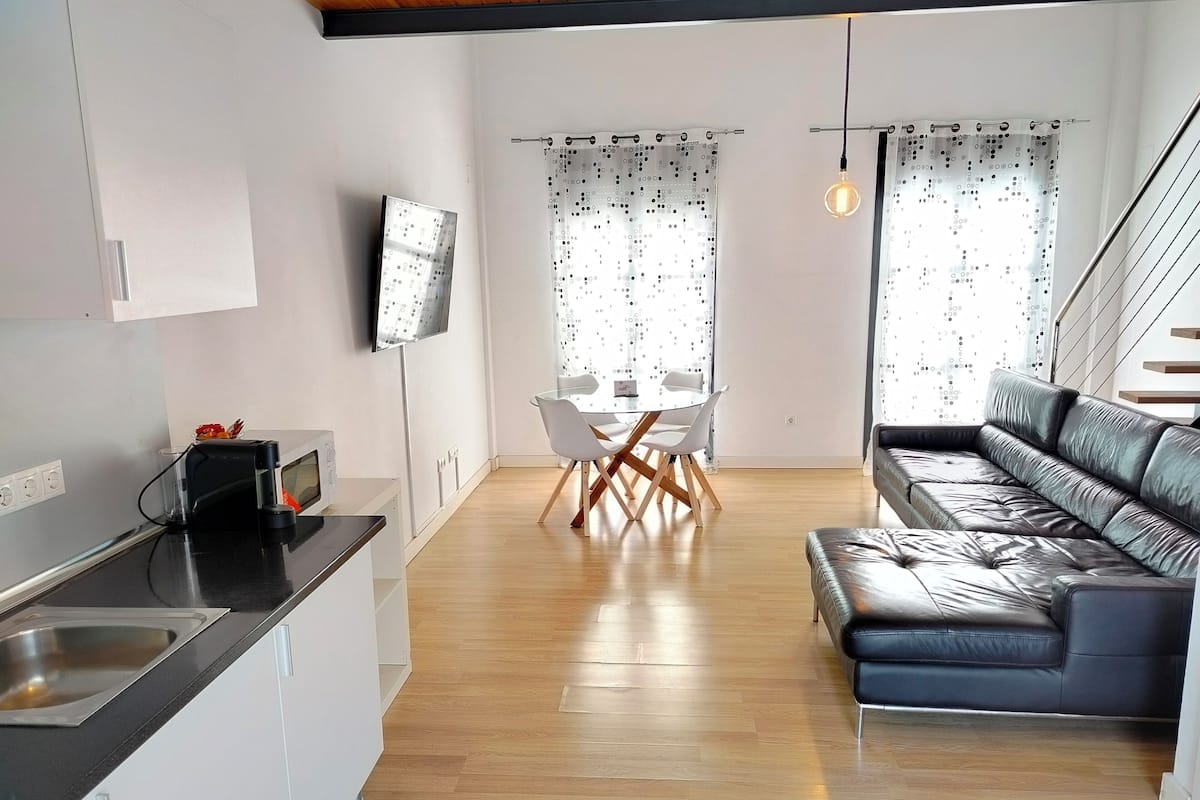 A modern kitchen with a black countertop and stainless steel sink, a dining area with a glass table and white chairs, and a dark leather sofa.