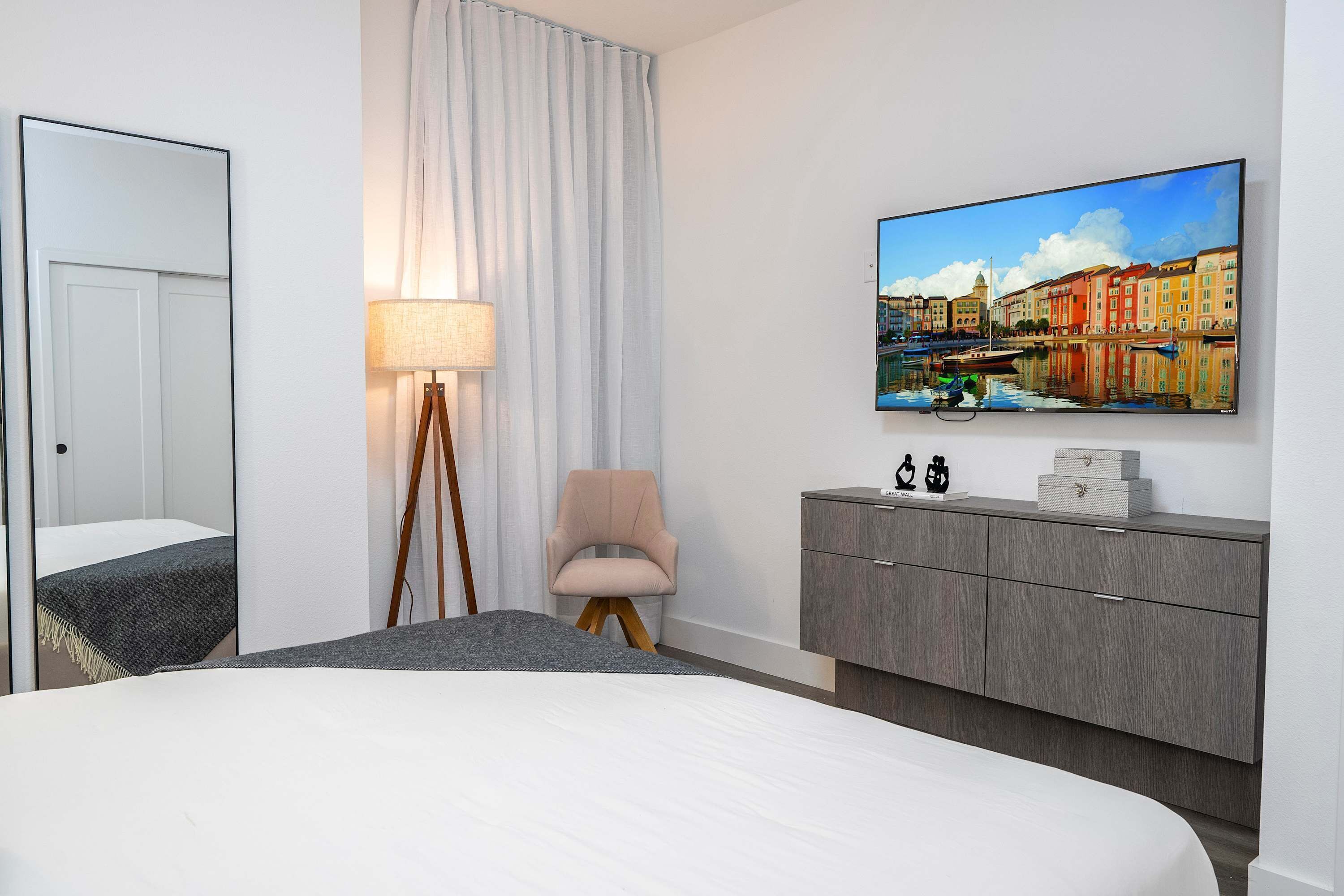 Une chambre d’hôtel moderne avec un grand lit, un téléviseur à écran plat fixé au mur, une commode grise, une lampe et une chaise.
