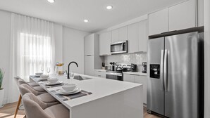 A modern kitchen with a central island, stainless steel appliances, and a dining area with chairs and a table set for four. - Doppio Luxury Home Themed Magic (Davenport)