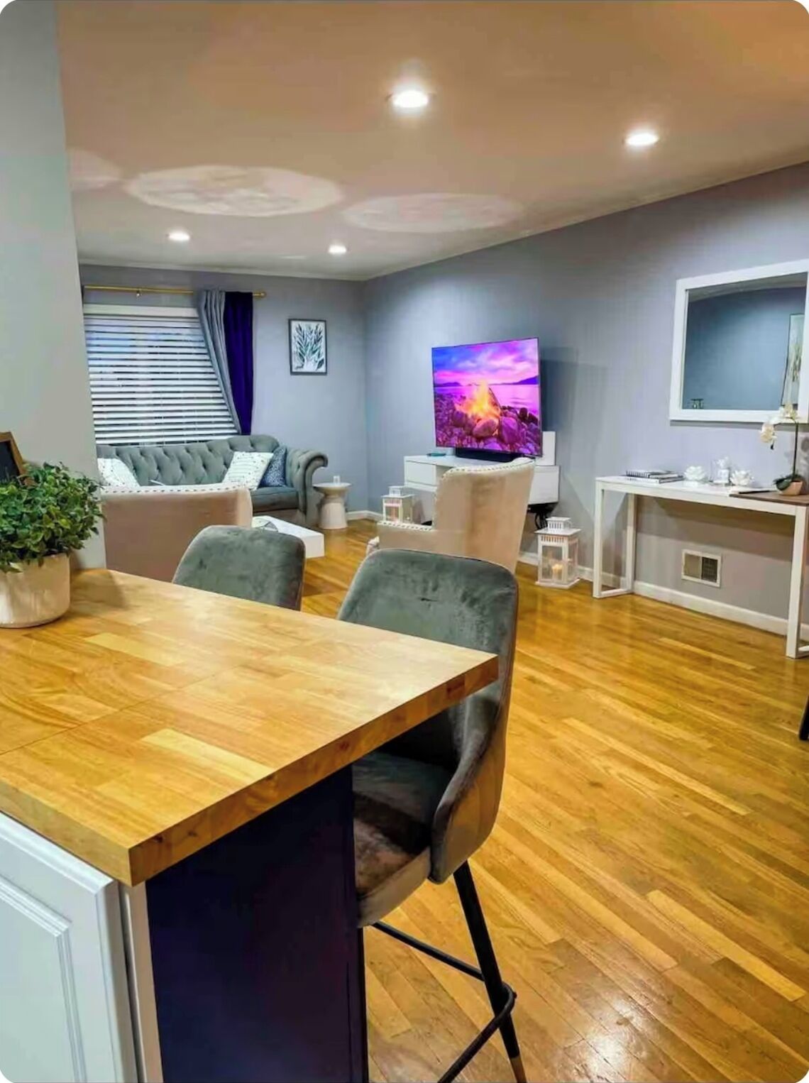 A modern living room with a wooden dining table, grey upholstered chairs, a grey sofa, a flat-screen TV, and a white console table.