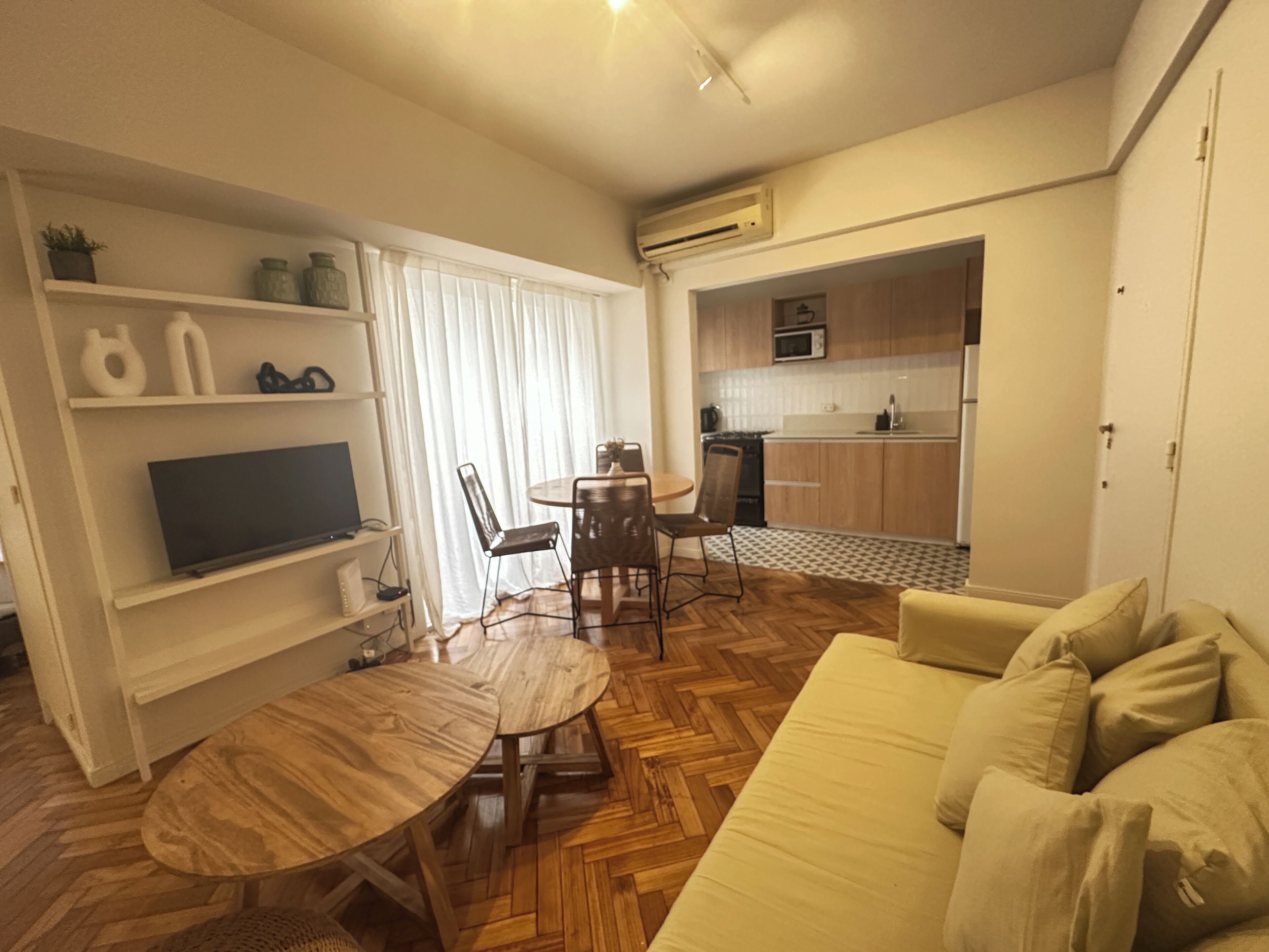 Een ruimtelijke woonkamer met een bank, een televisie op een plank, een eettafel met stoelen en een keuken met kastjes en apparatuur.