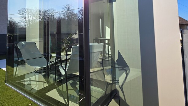 Une structure moderne en verre offrant une vue dégagée sur un espace de détente intérieur, comprenant des chaises et une table.