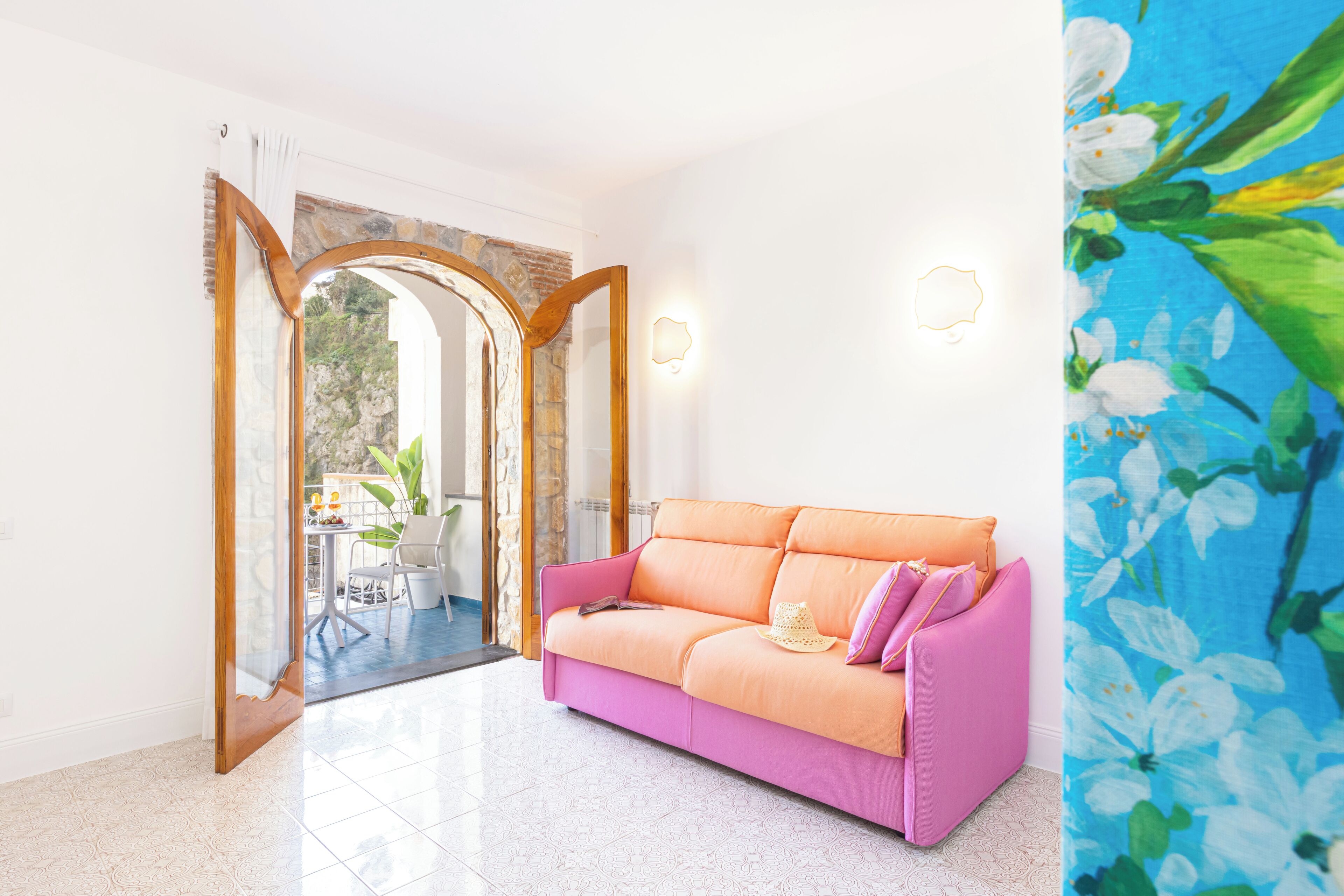 A pink sofa with a woven basket and a hat, a stone archway, and a view of an outdoor patio with a table and chairs.