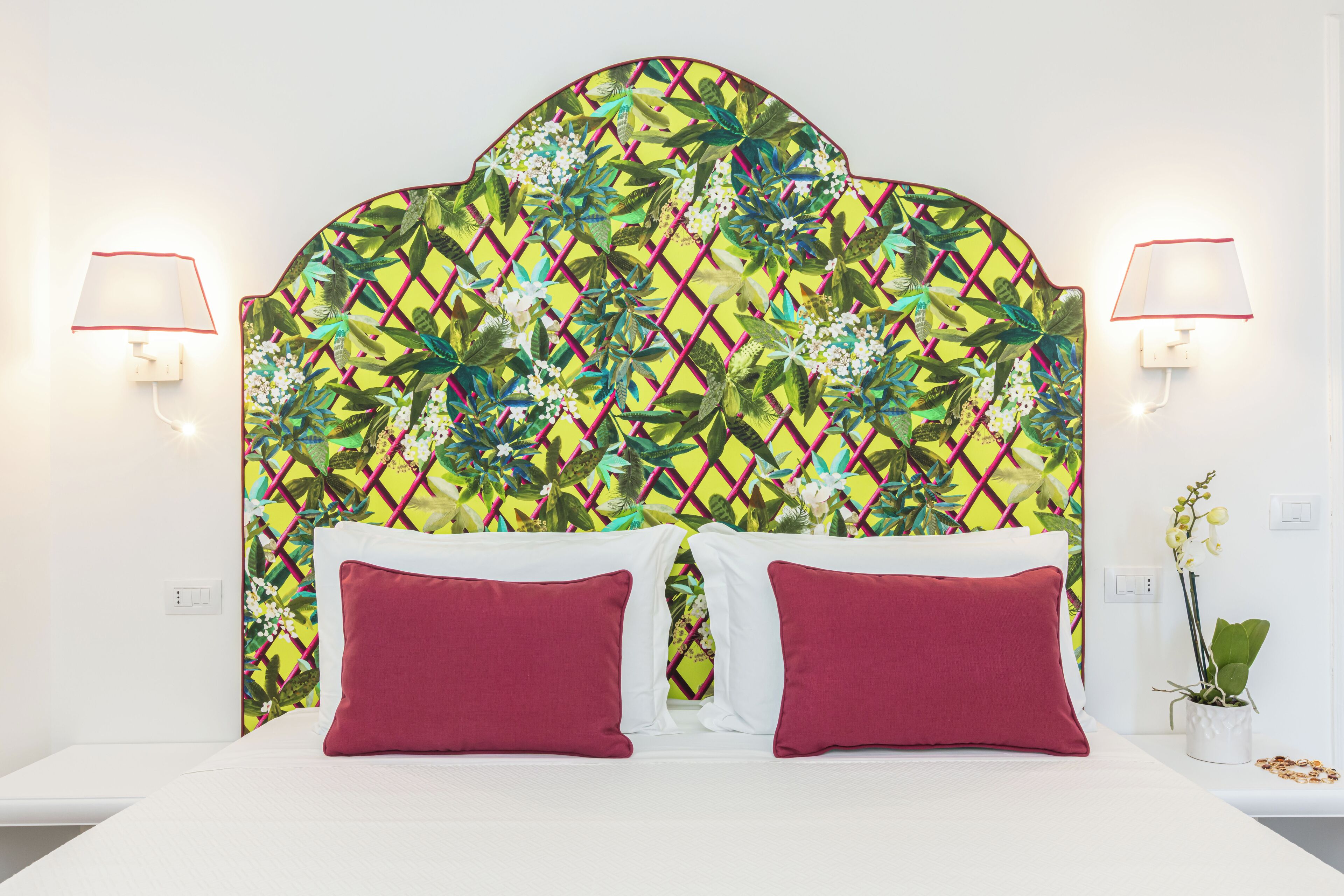 A hotel room with a bed, two red pillows, a floral-patterned headboard, and two wall-mounted lamps.