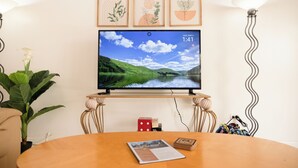 A room with a TV on a stand, a table with a notebook and dice, a plant, and a lamp. - Bayview Paradise Palms 2-bedroom Condo (Nassau)