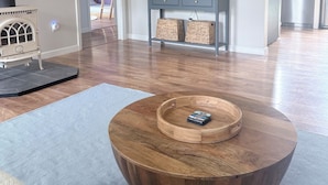 A living room with a large wooden coffee table, a flat-screen TV mounted on the wall, a kitchen area with modern appliances, and a visible door leading to another room. - The Central Ranch— minutes to Acadia & Coastal Maine (Ellsworth)