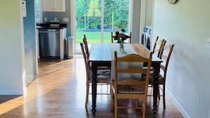 A dining area with a wooden table and chairs, a kitchen in the background, and a view of the outdoors. - The Central Ranch— minutes to Acadia & Coastal Maine (Ellsworth)