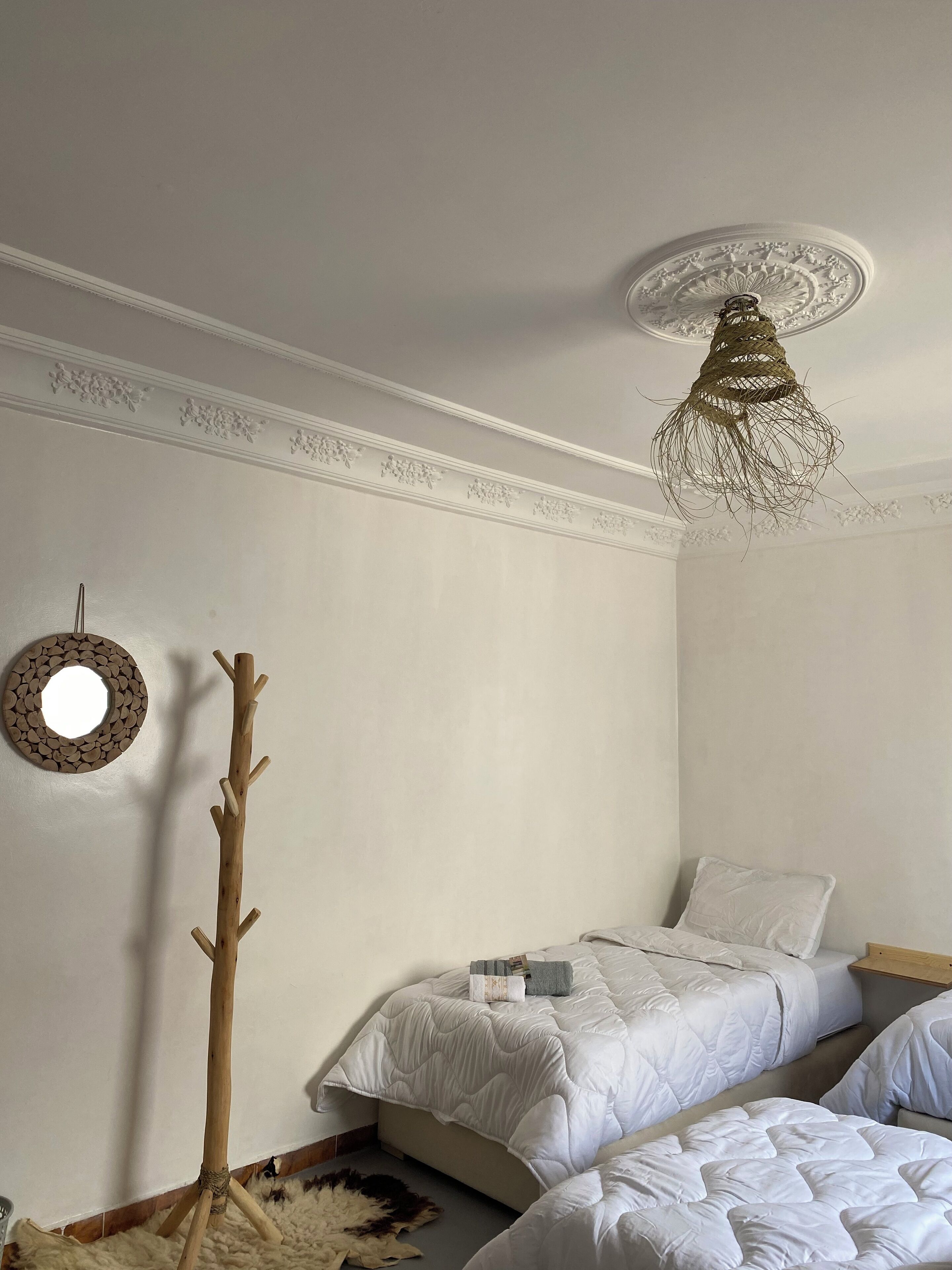 A bedroom with two beds, a wooden coat rack, a decorative ceiling light, and a circular mirror on the wall.