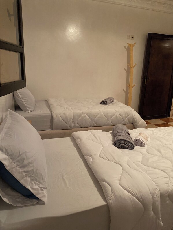Una camera d'albergo con due letti singoli, ognuno con piumino e cuscini bianchi. Si nota un appendiabiti in legno appoggiato al muro e una porta in legno scuro.