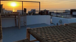 A rooftop terrace with a view of the sunset over the sea, featuring wicker furniture and a metal railing. - TITRYTE SURF HOUSE (Agadir)