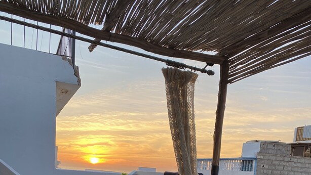 A balcony with a white railing, a patterned sofa, and a view of the sunset. - TITRYTE SURF HOUSE (Agadir)