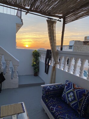 A balcony with a white railing, a patterned sofa, and a view of the sunset. - TITRYTE SURF HOUSE (Agadir)