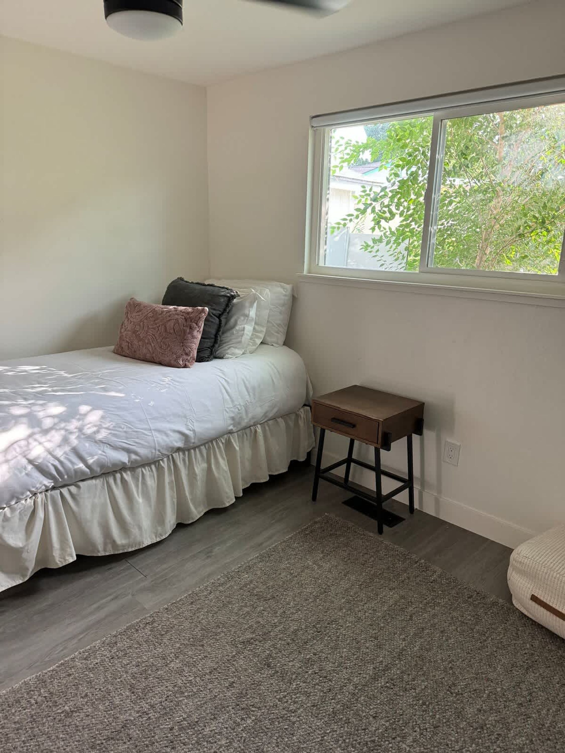 Un dormitorio con cama, mesita de noche, ventana y vistas al verdor exterior.
