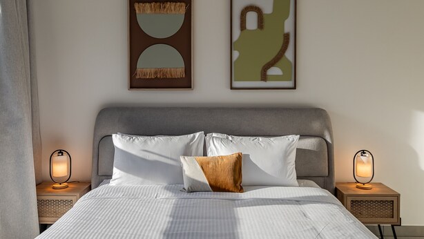 A neatly made bed with white linens and a brown pillow, flanked by two bedside tables with lamps, and two framed abstract artworks on the wall. - Socio Tower 1 206 Dubai Hills (Dubai)