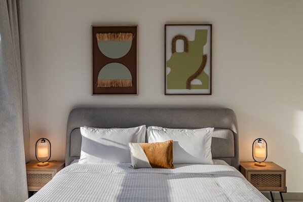 A neatly made bed with white linens and a brown pillow, flanked by two bedside tables with lamps, and two framed abstract artworks on the wall. - Socio Tower 1 206 Dubai Hills (Dubai)