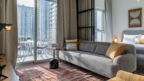 A modern hotel room with a large sliding glass door to a balcony, a grey sofa, a bed with white bedding, and a patterned rug. - Socio Tower 1 206 Dubai Hills (Dubai)