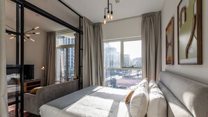 A modern bedroom with a large bed, a glass partition, a city view, and a ceiling light fixture. - Socio Tower 1 206 Dubai Hills (Dubai)