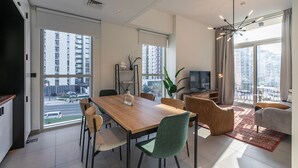 A modern living room with a wooden dining table, comfortable seating, a television, and large windows offering city views. - Socio Tower 1 206 Dubai Hills (Dubai)