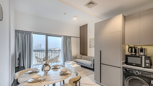 A modern kitchen with a built-in washer and dryer, a dining area with a round table and chairs, and a living area with a sofa and a view of the city. - MAG Building 920 711 (Dubai)