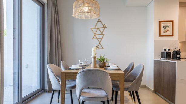 A modern dining area with a wooden table, grey upholstered chairs, a hanging light fixture, and a geometric wall decoration. - Tilal AL Ghaf 245 in Dubai (Dubai)