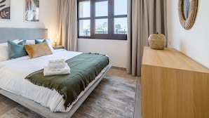 Quarto de hotel com uma cama grande, vista da cidade através de uma janela e mesa de cabeceira de madeira com abajur.