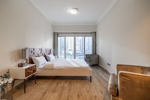 Ein modernes Schlafzimmer mit einem großen Bett, einem Nachttisch, einem Sessel und Blick auf hohe Gebäude durch große Fenster.