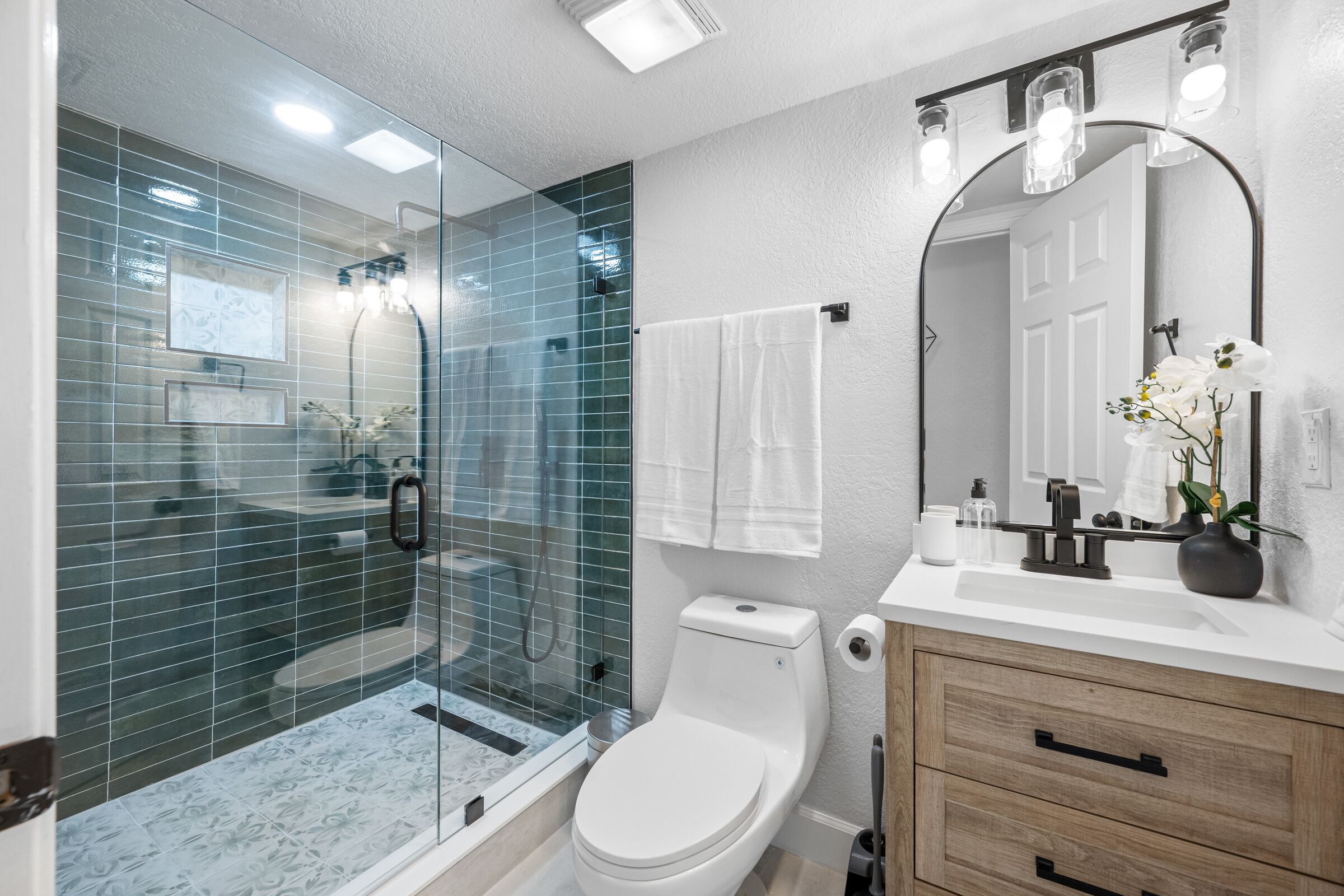 Un baño moderno con ducha de vidrio, inodoro blanco, tocador de madera con espejo y lavabo blanco.