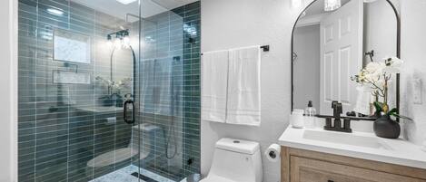 Un baño moderno con ducha de vidrio, inodoro blanco, tocador de madera con espejo y lavabo blanco.