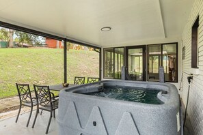A hot tub in a covered outdoor area with a table and chairs, large windows, and a grassy yard.