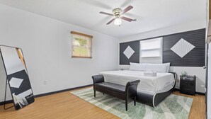 Une chambre à coucher comprenant un lit, une table de chevet, une chaise, un ventilateur de plafond et une fenêtre avec des stores.