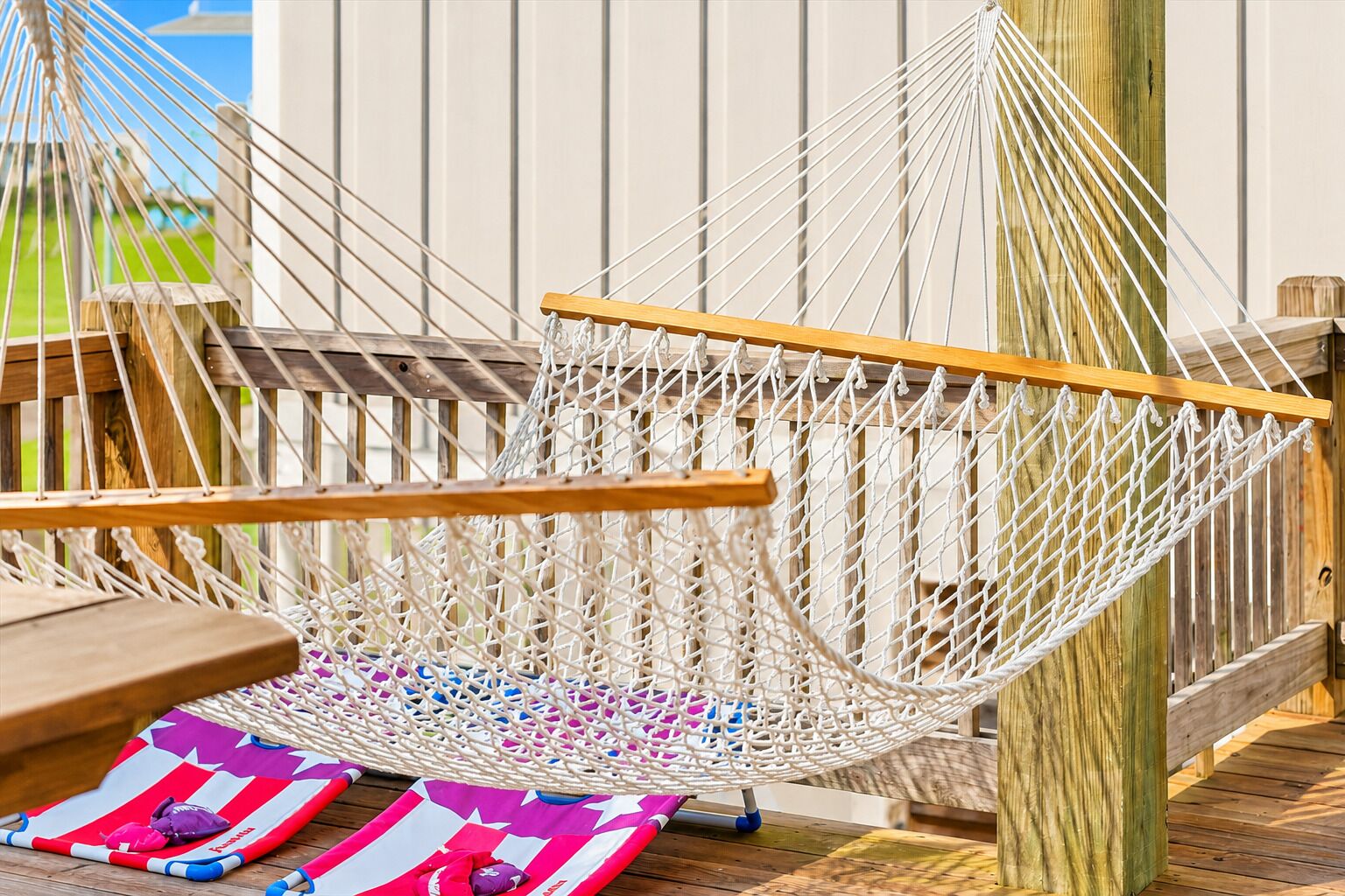 A hammock chair with a wooden frame and white netting, placed on a wooden deck with a wooden fence in the background.