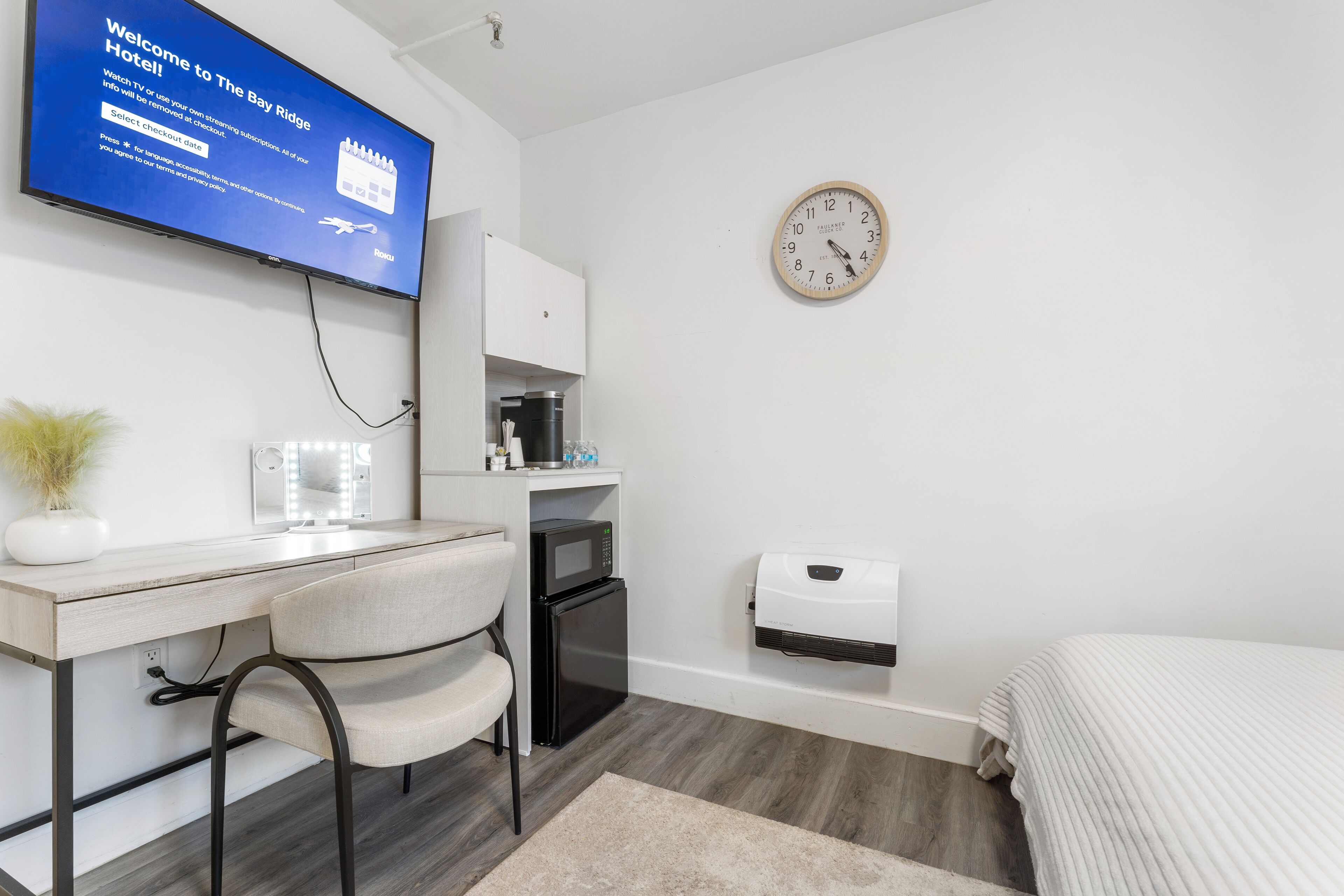 Ein modernes Hotelzimmer mit Flachbildfernseher, Schreibtisch mit Stuhl, Mikrowelle, kleinem Kühlschrank, einer Uhr und einem Bett.