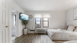 A bedroom with a bed, a radiator, a TV, and a window with blinds. - Bay Ridge ApartHotel (Brooklyn)
