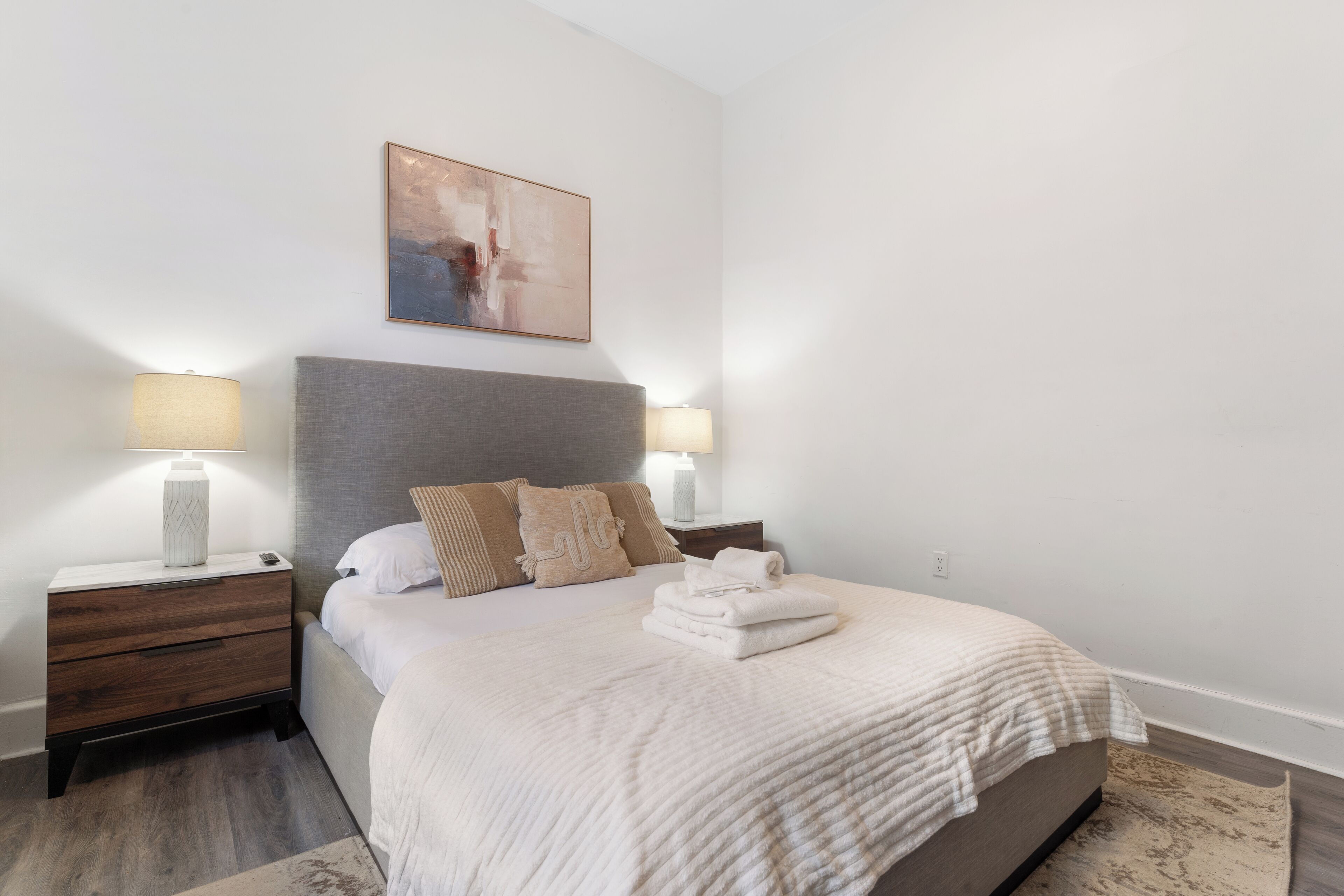 A neatly arranged bedroom with a bed, two bedside tables, a nightstand, and a painting on the wall.