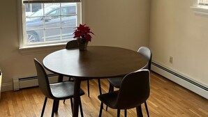 Une pièce avec une table ronde, quatre chaises, une plante en pot et deux fenêtres équipées de stores.